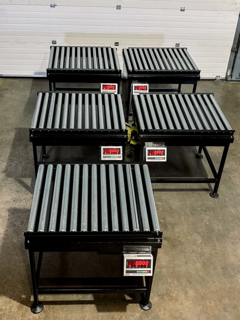Five weighing machines in a row with conveyor rollers and digital weighing display